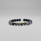 Pulsera elástica para hombre elaborada con cuentas de piedra volcánica negra, ojo de tigre dorado, soladita y piedras iridiscentes multicolor, con separadores metálicos en acero inoxidable, sobre fondo neutro gris claro.