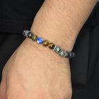 Pulsera elástica para hombre elaborada con cuentas de piedra volcánica negra, ojo de tigre dorado, soladita y piedras iridiscentes multicolor, con separadores metálicos en acero inoxidable, sobre fondo neutro gris claro. mano modelando